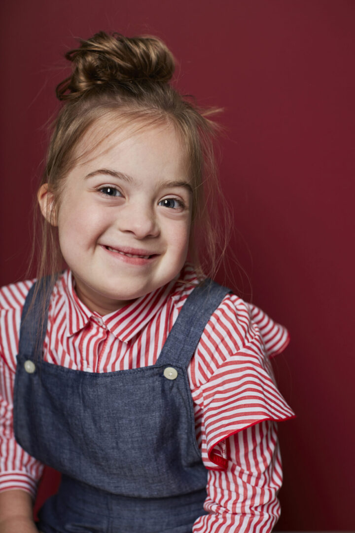 A young girl smiling for the camera in front of a red wall.
